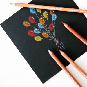 A black piece of paper with a colorful pastel drawing of balloons in red, blue, and yellow. Four MultiPastel® chalk pencils are placed around the paper: one yellow, one red, one orange, and one with a blue tip.