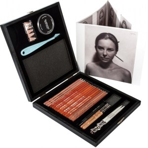 A black box opened to reveal a set of drawing tools, including pencils, a pencil sharpener, an eraser, and blending tools. Accompanying the set is a booklet featuring a black and white portrait of a woman on the cover.
