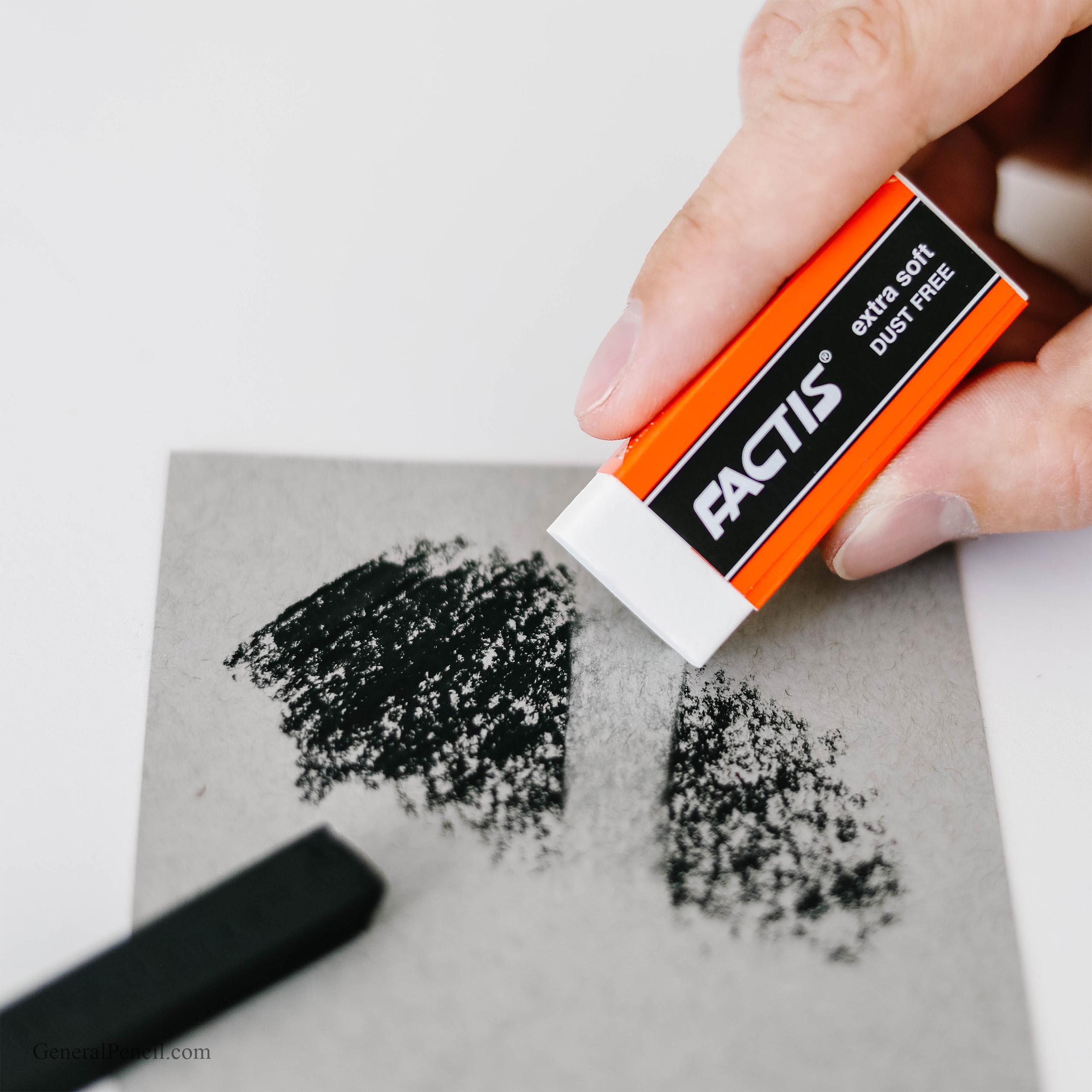 A hand uses a Factis® Extra Soft White Vinyl Eraser to remove charcoal marks from grey paper, with a compressed charcoal stick nearby.