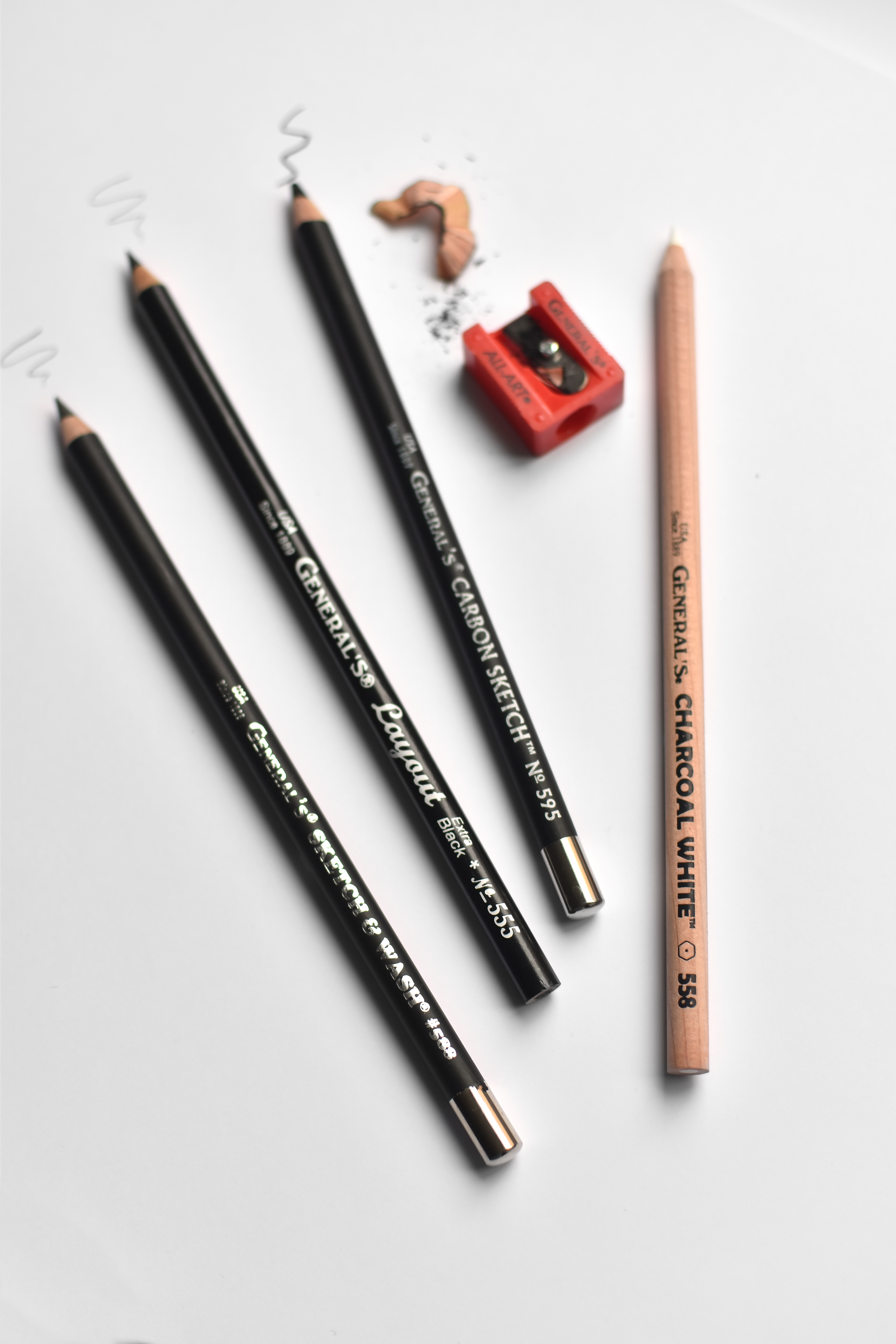 Four pencils from the Hand Lettering Pencil Set, 14 pieces, are neatly placed on a white surface: three being black carbon sketch pencils and one a white charcoal pencil. Accompanying them is a red pencil sharpener and some shavings, with small pencil doodles visible nearby.