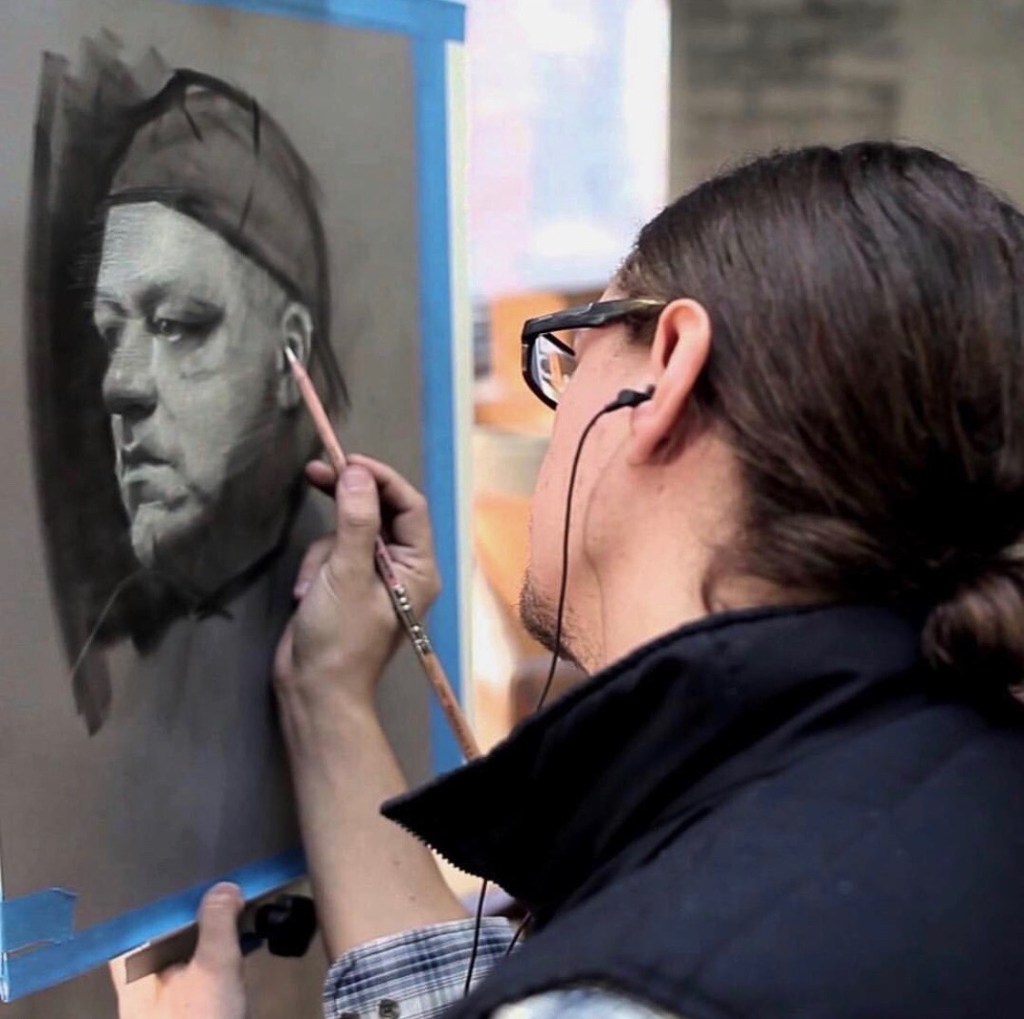 Artist David Kassan draws John with General's® Charcoal White® Pencil. David is looking at the drawing while wearing a blue and white plaid shirt with blue vest and earbuds in. On the end of the Charcoal White® Pencil is a Miser® pencil extender.