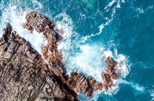 Aerial view of rocky cliffs jutting into the turquoise ocean. White waves crash against the rocks, creating frothy splashes and foam. The scene captures the rugged coastline's dynamic interaction with the sea.