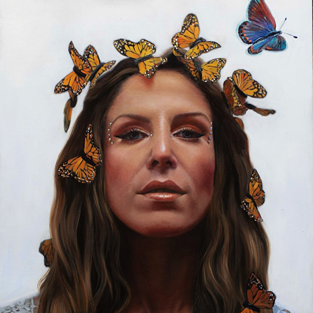 Artist Shana Levenson with long hair is adorned with orange butterflies on her head and face. She has a serene expression and subtle makeup with white dots around her eyes. A single blue butterfly hovers near her head against a plain white background.