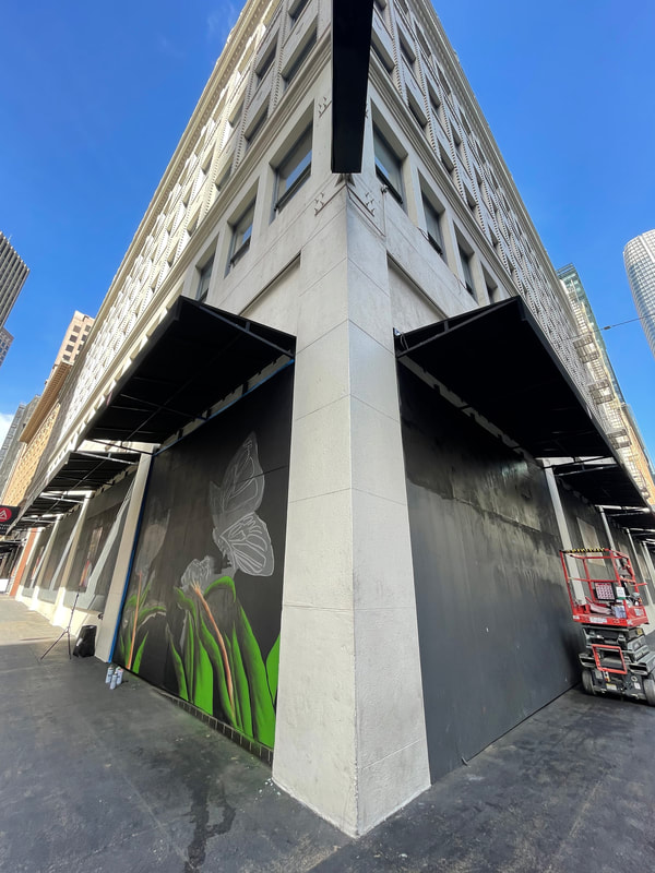 This is an image of the corner of a multistory city building looking up from a low angle. The first floor sides of each corner has the beginnings of a mural. A red and grey scissor lift is on the right side of the image. There is clear blue skies and at all city building visible on both sides of the image.
