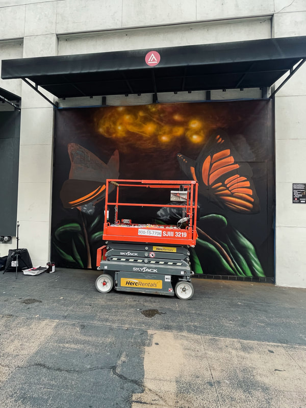 Side of a multistory city building with a black awning with the red and white Academy of Art University logo on it. Below the awning is an in-progress monarch butterfly mural with one butterfly and green outlines below it. There is a red and grey scissor lift in front of it on the sidewalk. A tripod and black bag are on the left side of the mural.