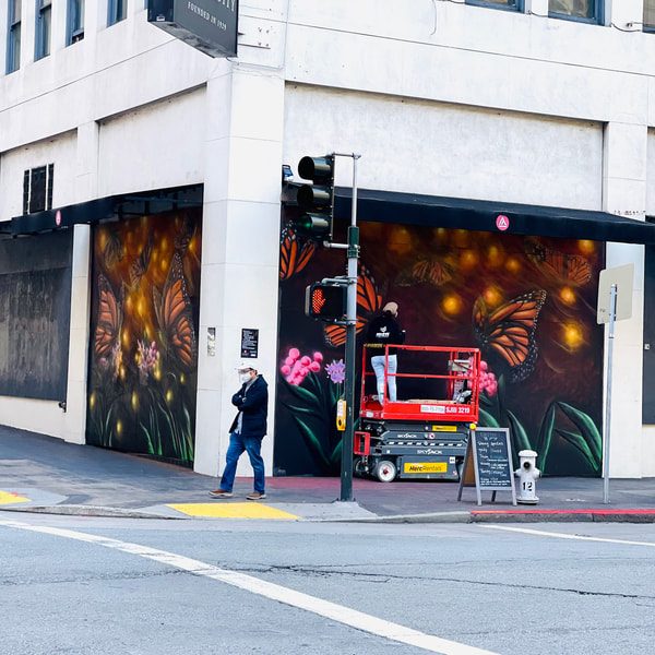Corner of a white multistory building with a monarch butterfly mural painted on both sides of the corner. In front of the mural on one side is a red and grey scissor lift. There is a sandwich board sign and fire hydrant in front of that. In front of the building on the street corner is a man wearing a white hat, black jacket, blue jeans, and a face mask. At the corner there is a crosswalk.