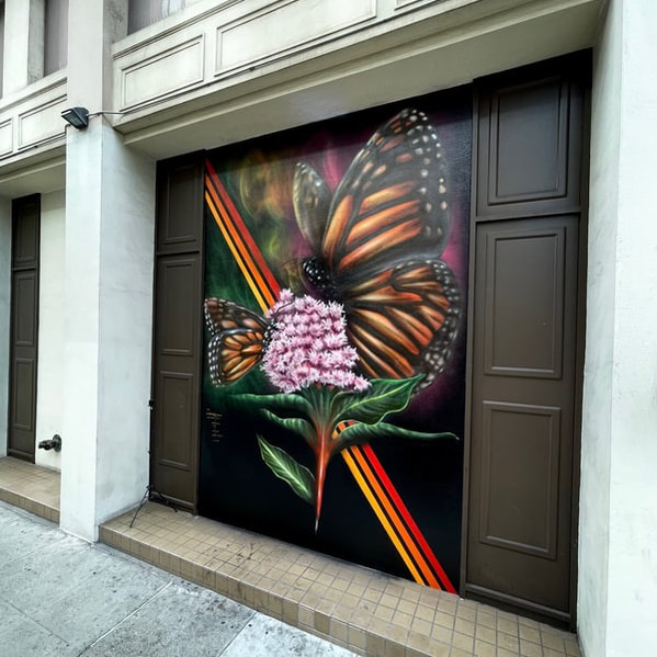 A finished monarch butterfly mural with one large and one small orange, white, and black monarch butterfly. The butterflies are sitting atop a plant with a red stem, green leaves, and pink and white flowers. The are stripes of red, orange, and yellow bisecting the background from the top left to the bottom right of the mural. The large butterfly in the top right of the image is surround by a pinkish purple background with swirls of yellow on the left side. The small butterfly on the right side of the image has a wispy green background behind it. The mural is surrounded on either side by three vertical panels painted dark brown with rectangular insets in the same color. At the base of the mural is a one tile high and four and a half tile deep ledge. The tiles are dirty and tan in color. The building this are is set in is white with three horizontal panels with white rectangular insets above it.