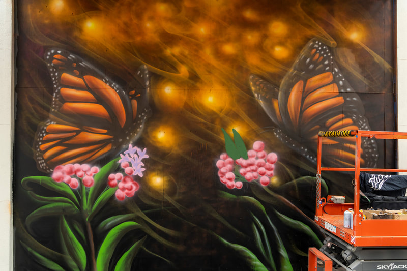 Image of an in-progress monarch butterfly mural with two monarch butterflies each perched atop a green leafy plant with red berries and pink flowers. The background is orange with "glowing" spots. An orange and grey scissor lift is partially in front of the mural.