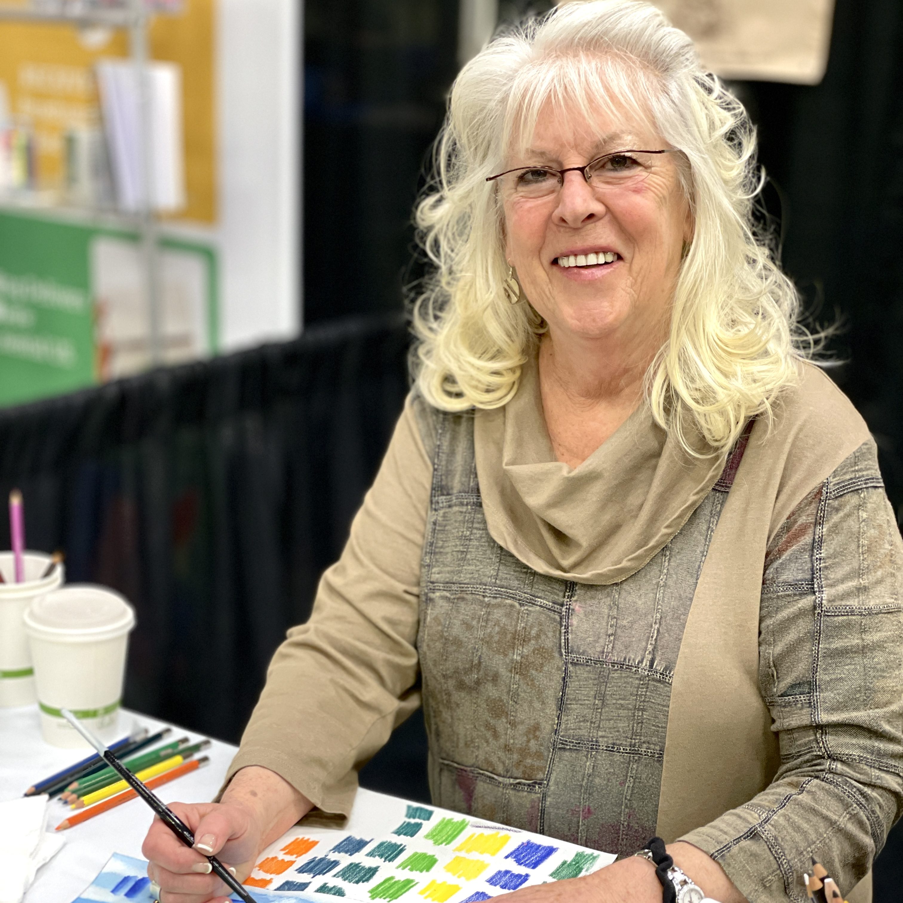 Artist Kathi Hanson, with long, wavy silver hair smiles while painting with watercolor pencils at a table. Art supplies and color swatches are visible in front of her. She wears glasses and a tan patterned top.