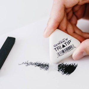 A hand holds a white, triangular Factis® Tri-Tip™ Eraser, partially erasing a dark charcoal smudge on white paper, with a compressed charcoal stick lying nearby.