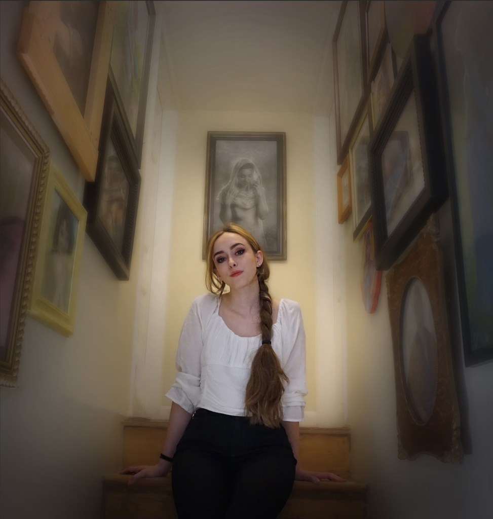 A woman with long brown hair in a braid sits on stairs, wearing a white blouse and black pants, surrounded by framed art and portraits on the narrow stairwell walls. A black-and-white charcoal drawing portrait hangs above the stairs.