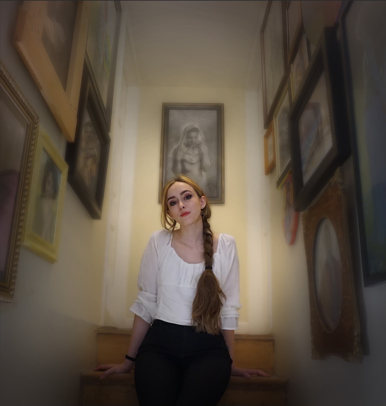 A woman with long brown hair in a braid sits on stairs, wearing a white blouse and black pants, surrounded by framed art and portraits on the narrow stairwell walls. A black-and-white charcoal drawing portrait hangs above the stairs.