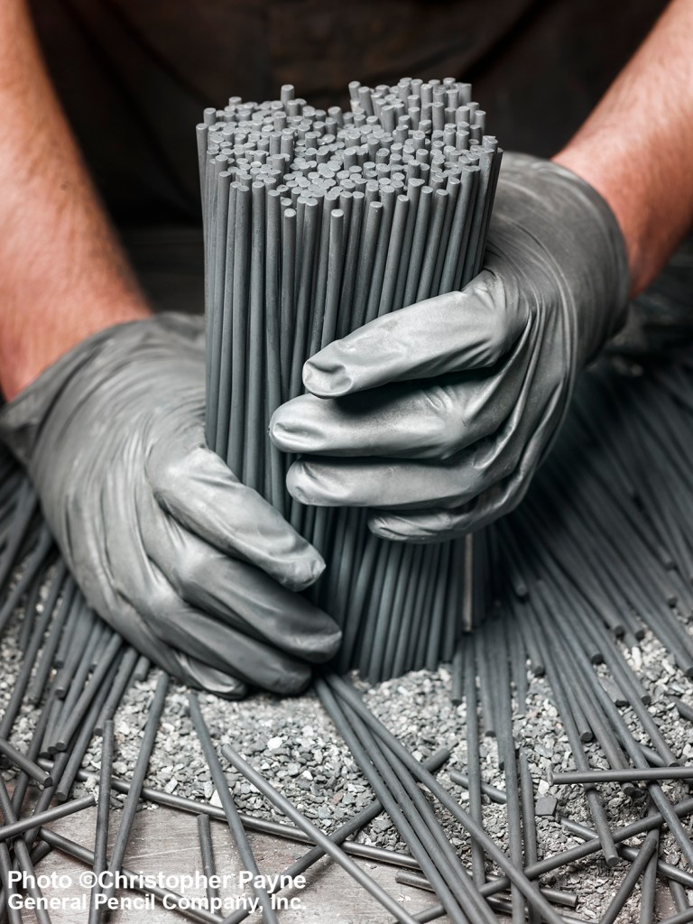 A person wearing gray gloves holds a large bundle of graphite rods, surrounded by scattered rods and graphite fragments on a work surface.