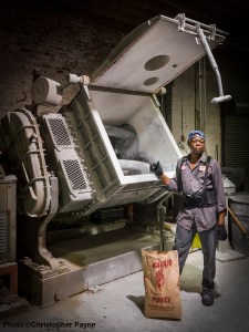 A worker in a uniform and safety gear stands beside an industrial mixer filled with white powder, holding a bag labeled "Pumice." The scene is set in a factory with exposed brick walls and machinery.