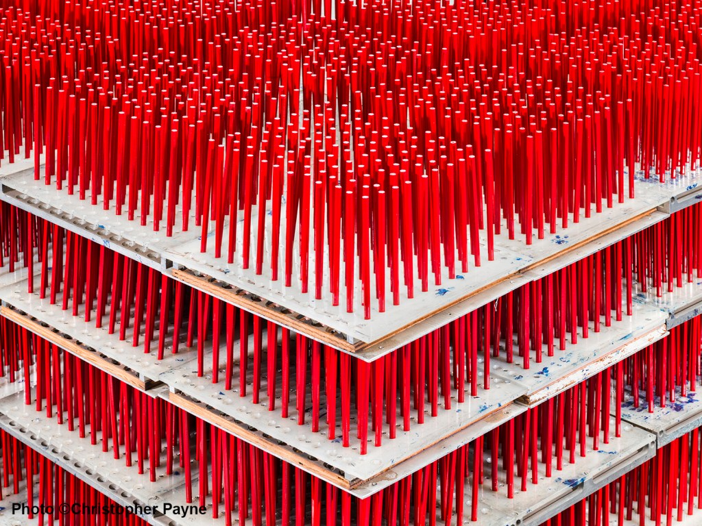 Hundreds of red pencils stand upright on stacked trays, forming a dense, organized pattern. The trays are layered, creating a geometric, abstract visual effect. Photo by Christopher Payne.