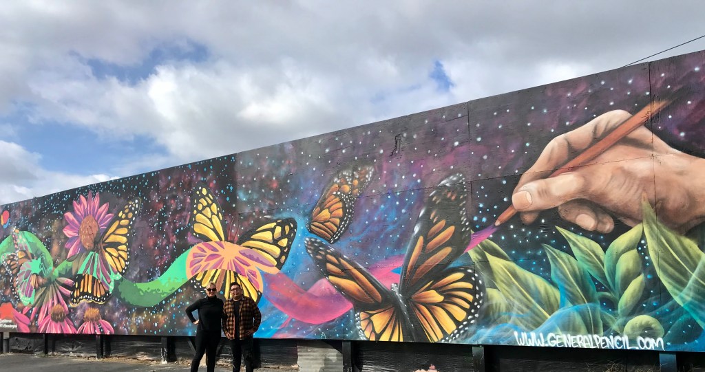 A colorful mural depicts a hand painting butterflies, flowers, and leaves against a starry background. Two people stand in front of the mural, which covers a long wall under a partly cloudy sky.