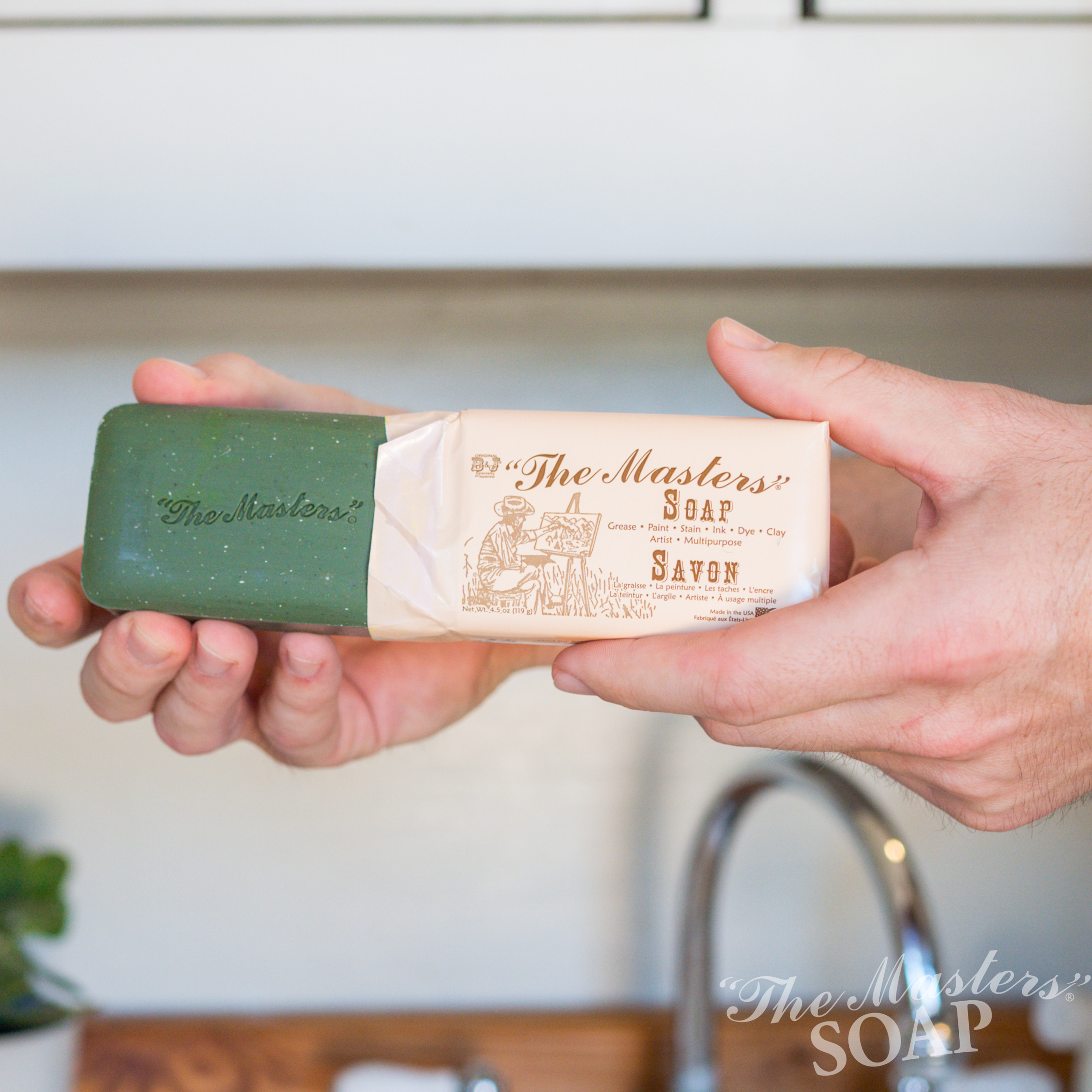 A person holds a green bar of "The Master's Soap," partially unwrapped from its beige paper packaging, standing in a bright kitchen with a sink and small plant in the background.