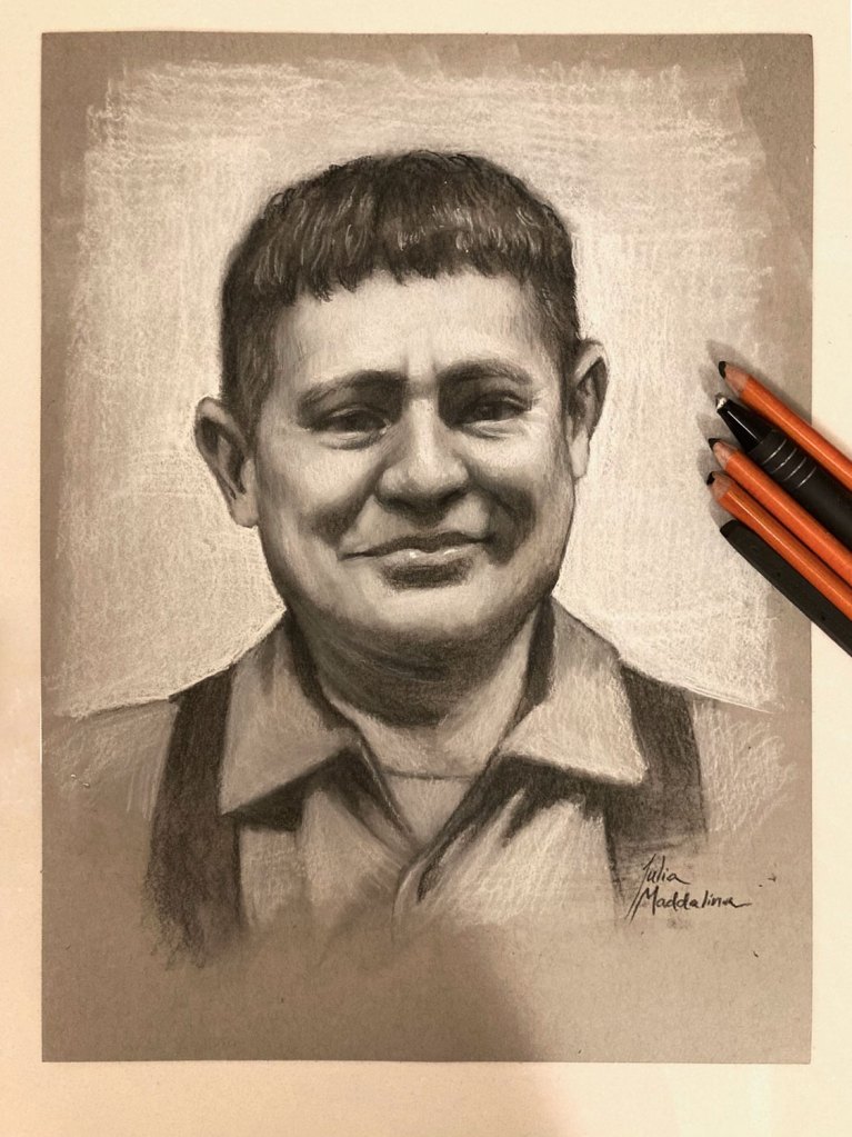 A realistic pencil portrait of a smiling man on gray paper, with three pencils—black, orange, and red—placed to the right of the drawing. The artist’s signature is visible in the lower right corner.